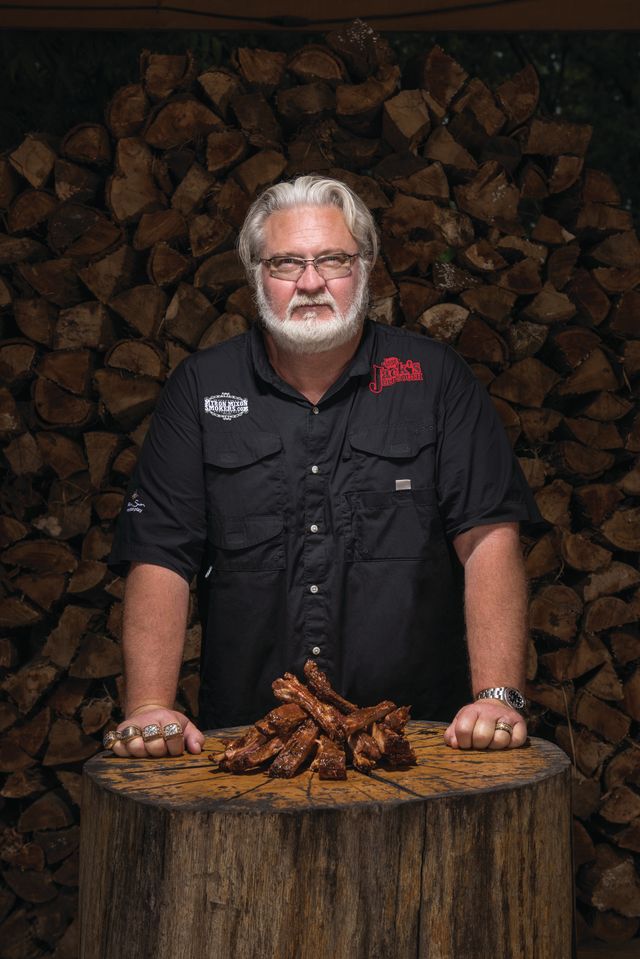 Myron Mixon standing at a chopping block with smoked ribs in front of a wood pile